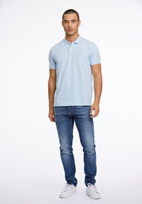 Polo azzurro chiaro con maniche corte, colletto a bottoni e logo discreto sul petto, abbinato a jeans blu slim-fit e sneakers bianche.