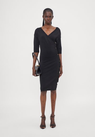 Schwarzes, figurbetontes Kleid mit Dreiviertelärmeln, V-Ausschnitt, Knopfdetails auf einer Seite und einem strukturierten Stoff. Modell hält eine kleine schwarze Handtasche.