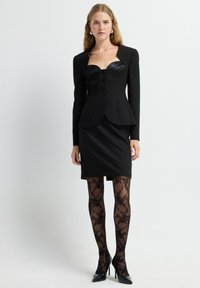 Blazer negro con detalles de satén, cuerpo ajustado, seis botones, combinado con una falda negra y medias negras transparentes con patrón. Los tacones altos completan el look.