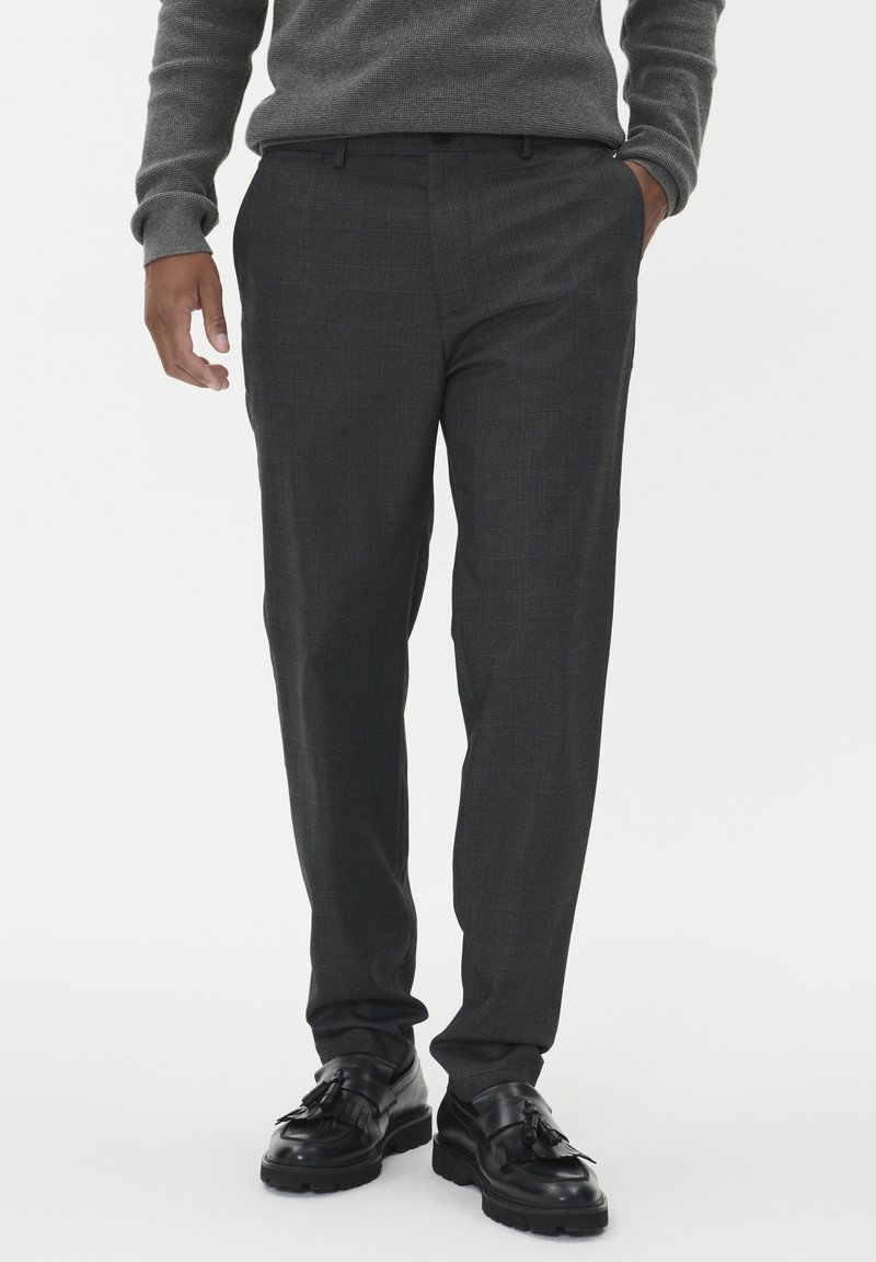 Moški nosi sive črtaste oblečne hlače, črne loafers z resicami ter sivi dolgi pleteni pulover, z rokami delno v žepih.