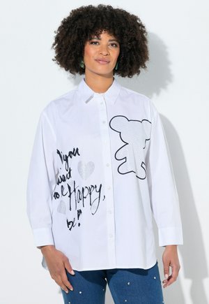 Kobieta o kręconych włosach, ubrana w białą koszulę zapinaną na guziki z czarnymi szkicami, napisem "Chcesz być szczęśliwy, bądź!" oraz wzorem zarysu niedźwiedzia.