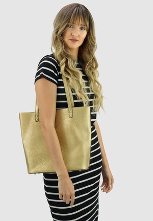 Vrouw met lang golvend haar, gekleed in een zwart-wit gestreepte jurk, die een grote metallic gouden schoudertas draagt.
