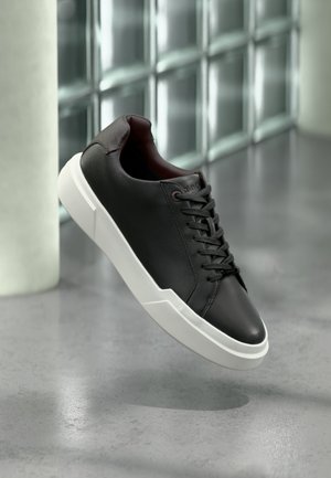 Baskets en cuir noir avec une semelle blanche épaisse, lacets noirs, flottant au-dessus d'un sol en béton gris près d'un mur en blocs de verre.