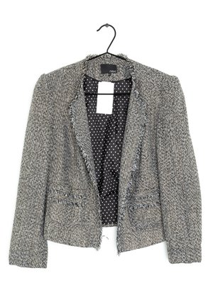 Blazer gris texturé pour femme avec bords effilochés et poches avant, doublure à pois noirs et blancs, suspendu sur un cintre noir.