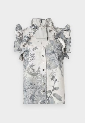 Femme portant une robe midi sans manches à fleurs avec volants, boutons et un ourlet asymétrique, associée à des talons noirs à bride à la cheville.