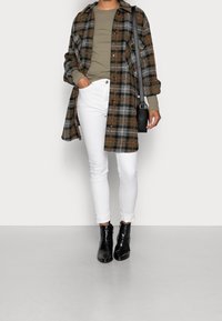 Person, die einen karierten braun-grauen Mantel, ein olivgrünes Oberteil, weiße Hosen und schwarze Stiefeletten trägt und eine schwarze Umhängetasche dabei hat.