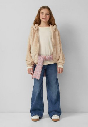 Lichtbeige fluffy ritsvest, crèmekleurig top, roze met patroon sjaalriem, wijdvallende blauwe jeans en witte sneakers met beige zolen.