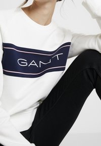 Biała bluza z okrągłym dekoltem z granatowym i różowym prostokątnym paskiem z logo "GANT". Model siedzący, w czarnych spodniach, widok pod kątem. Gładka struktura tkaniny.