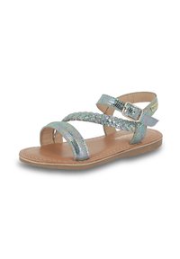 Sandal plat avec une semelle marron clair, des brides tressées iridescentes et une fermeture à boucle. Présente une texture lisse et un design minimaliste.