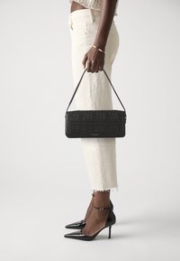 Borsa a tracolla nera intrecciata, con forma rettangolare liscia, rifiniture in pelle e tracolla, tenuta da una persona che indossa pantaloni beige e tacchi neri.