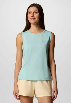 Donna che indossa una canottiera azzurro chiaro e pantaloncini giallo chiaro, in piedi e guarda leggermente di lato con uno sfondo neutro.