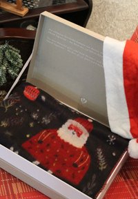 Schwarzer Schal mit einem gestrickten Santa-Design in roter Kleidung, präsentiert in einer weißen Box mit grauem Text. Begleitet von einer roten und weißen Santa-Mütze.