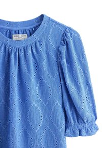 Blus i blått texturerat tyg med diamantformade hålmönster, rynkad halsringning och lätt puffade ärmar med volangprydda muddar.