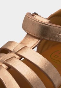 Ιμάντες σανδαλιών σε μεταλλικό ροζ χρυσό με κλεισίματα αστραγάλου με velcro και μαλακό καφέ πάτο με εμφανή σήμανση.