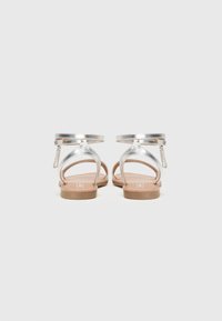 Zilverkleurige metalen sandalen met een platte zool, voorzien van dunne bandjes en een decoratieve kettingaccent aan de achterkant voor extra detail.