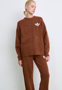 Brauner Strickpullover und passende Hose mit gerippten Bündchen. Der Pullover hat ein weißes Logo auf der Brust. Weiche Textur, lockere Passform.