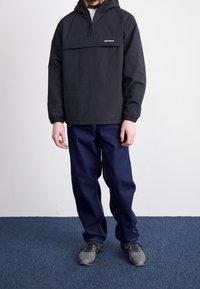 Persona con un anorak negro con capucha, jeans azul oscuro y zapatillas grises de pie sobre una alfombra azul contra una pared blanca.