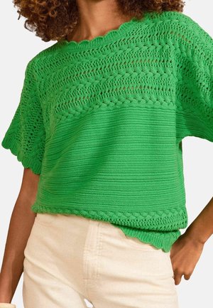 Kvinde iført lys grøn hæklede kortærmede top med bølgekant og cremefarvede bukser, poserer med hånden på hoften.