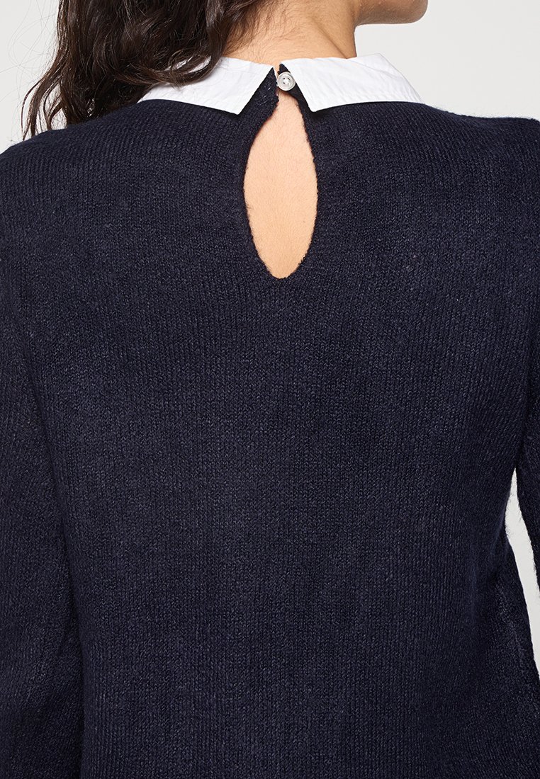 Pull marine bleu avec un tricot texturé, col rond, et un détail en trou de serrure à l'arrière, superposé sur une chemise blanche à col.