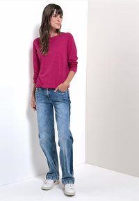 Fuchsias långärmad topp i kombination med ljusblå raka jeans och vita sneakers, stående mot en neutral bakgrund.