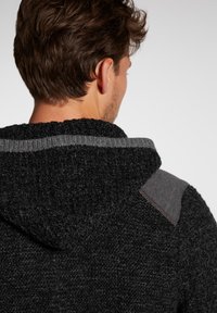 Texturierte schwarze Strick-Hoodie mit einem grauen Streifen am Halsausschnitt und grauen Akzenten an der Schulter, ausgestattet mit einer geräumigen Kapuze und einem lockeren Schnitt.