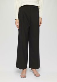 Schwarze weit geschnittene Hose mit glatter Textur, vorderen Falten und seitlichen Taschen. Kombiniert mit silbernen Schuhen mit offenem Zehenbereich.