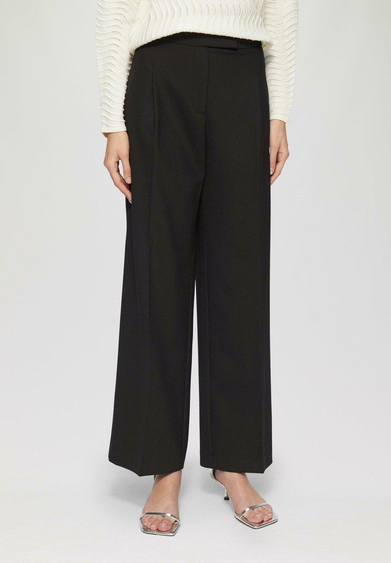 Schwarze weit geschnittene Hose mit glatter Textur, vorderen Falten und seitlichen Taschen. Kombiniert mit silbernen Schuhen mit offenem Zehenbereich.