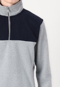Pullover in pile di colore grigio chiaro e blu navale, con collo alto, mezza zip e una superficie testurizzata. Blocco di colore distintivo nell'area del petto.