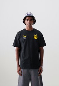 Svart kortermet T-skjorte med gul Dortmund-logo og Puma-merke. Modellen har på seg en rutete bøttehatt og grå cargo-shorts.