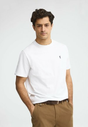 Hombre con cabello rizado oscuro, vistiendo una camiseta blanca de manga corta con un pequeño logo, pantalones marrones y un cinturón oscuro, con las manos en los bolsillos.