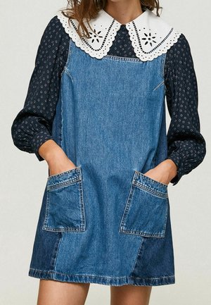 Jeansjurk met een knielange snit, grote voorzakken en een witte geborduurde kraag over een marineblauwe blouse met lange mouwen en een patroon.