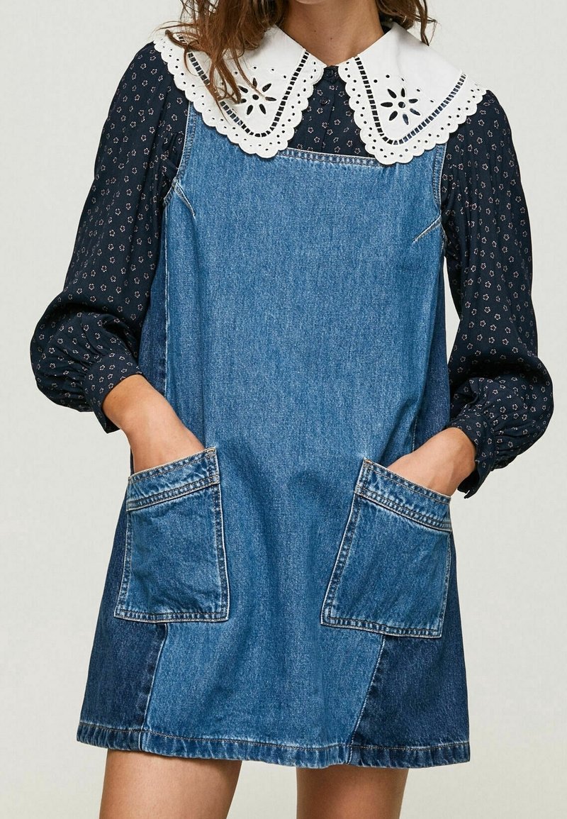 Jeanskleid mit knielangem Design, großen Fronttaschen und einem weißen bestickten Kragen über einer dunkelblau gemusterten Bluse mit langen Ärmeln.