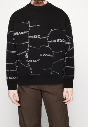 Person wearing black sweatshirt with repeated white "Armani Exchange" text and brown pants, standing against a plain background.