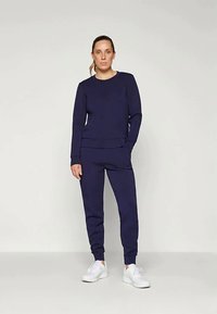 Námornícky modrý súprava mikiny a nohavíc z mäkkého materiálu, s okrúhlym výstrihom, elastickými manžetami a bočnými vreckami. Nosí sa s bielymi teniskami.