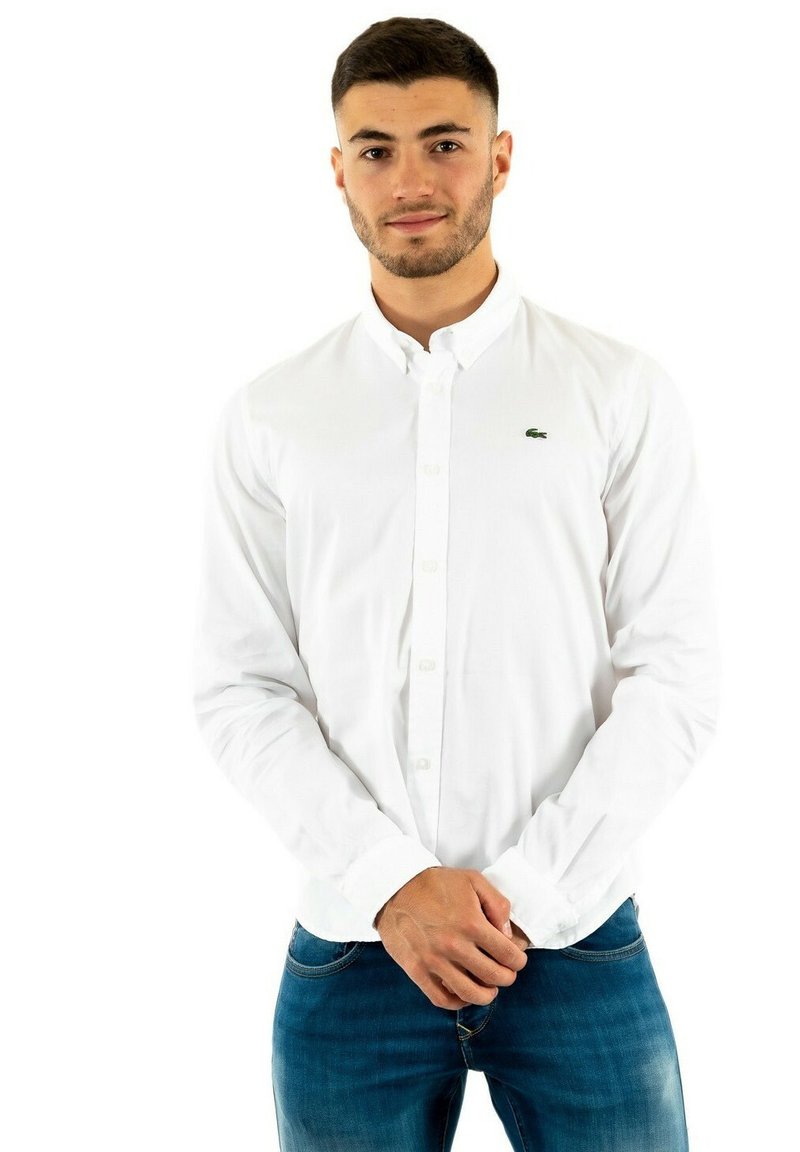 Chemise blanche à boutons en tissu coton, avec une coupe slim et un logo vert Lacoste sur la poitrine. Portée avec un jean en denim bleu.