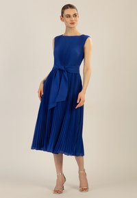 Faltenblaues Kleid mit Rundhalsausschnitt, tailliertem Oberteil und gebundenem Taillenband. Silberne Riemchenabsätze ergänzen das Outfit. Glatte Textur.