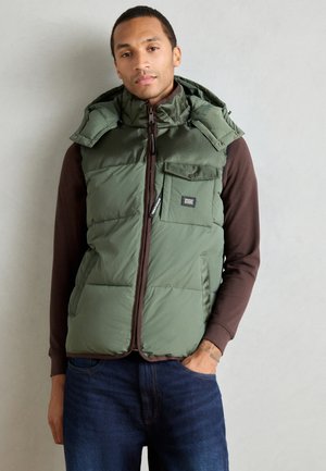 Green puffer vest with a zip front, two chest pockets, and a detachable hood, worn over a brown long-sleeve shirt. Smooth, insulated texture.