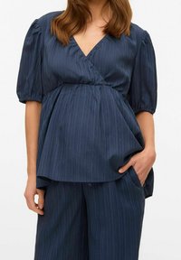 Blouse bleu marine texturée avec un col en V, des manches courtes bouffantes et une taille froncée. Présente de fines rayures verticales et une coupe ample.