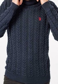 Mörkblå kabelstickad tröja med hög krage, ribbade ärmslut och fåll. Har en liten röd logotyp på vänster bröst. Texturerad mönster genom hela plagget.