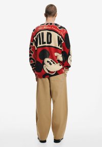 Red and beige oversized sweater featuring Mickey Mouse graphics, ribbed texture, and bold lettering. Paired with beige wide-leg trousers.