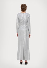 Ασημί φόρεμα μακρύ με μακριά μανίκια, εφαρμοστή σιλουέτα, λεπτομέρεια με φερμουάρ στην πλάτη και λεία, λαμπερή υφή που φτάνει μέχρι τους γίνες. Ορατές μαύρες γόβες.