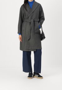 Grijze mantel van wolmix met wikkel en strikceintuur, brede mouwen, twee zakken, gecombineerd met een blauwe gebreide top, donkerblauwe wijde broekspijpen en zwarte sneakers.