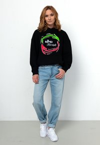 Schwarzer Hoodie mit mehrfarbigem Grafiklogo, kombiniert mit hellblauen Jeans und weißen Sneakers. Lässiger, lockerer Schnitt. Glatte Stofftextur.