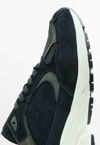 Scarpa atletica in suede blu scuro con dettagli in rete, suola intermedia bianca spessa e suola nera texture. Presenta un colletto imbottito e un design dei lacci.