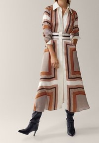 Robe chemise à motifs marron et blanc avec un col, des manches longues et une ceinture ; portée avec des bottes noires au genou. Texture de tissu lisse.