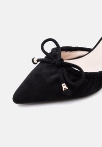 Chaussure plate noire à bout pointu avec côtés froncés et nœud décoratif orné d'accents métalliques argentés.