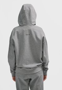 Osoba w jasnoszarej bluzie z kapturem i dopasowanych spodniach, widziana z tyłu, z napisem "F.U.T." wydrukowanym małymi czarnymi literami na plecach.