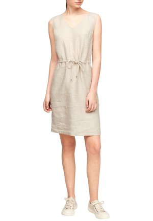 Kobieta ubrana w bezrękawnikową beżową lnianą sukienkę z wiązanym w talii sznurkiem oraz jasnobeżowe sportowe buty, stojąca na jednolitym białym tle.