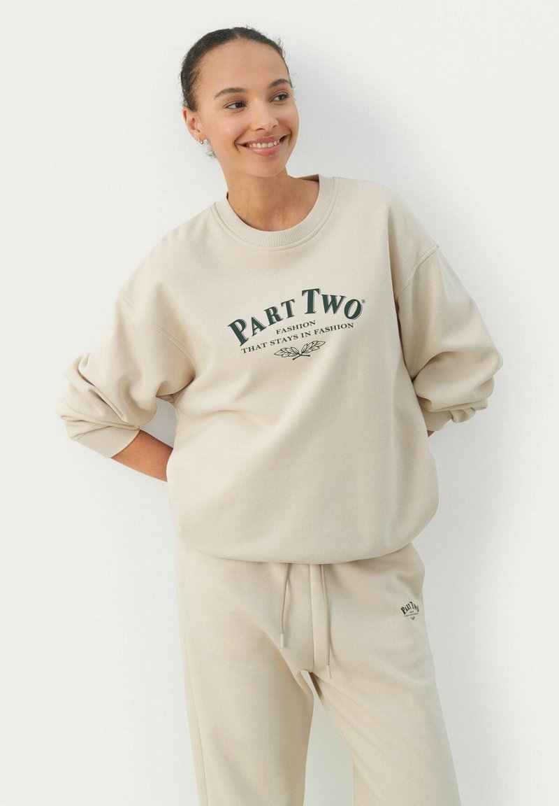 Felpa beige con logo "Part Two" in verde scuro, vestibilità ampia, maniche a cuffia, abbinata a pantaloni della tuta beige coordinati, con cintura regolabile.