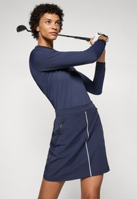 Námornícky modrý športový top s dlhým rukávom v kombinácii s rovnakou sukňou s bočnou vreckom na zips a bielymi bočnými detailami. Hladká, ľahká tkanina navrhnutá na pohyb.